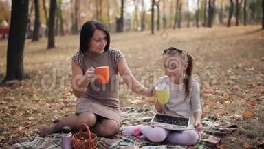一对可爱的母女正在公园里坐在格子布上喝茶。 小女孩在黑板上画着蜂